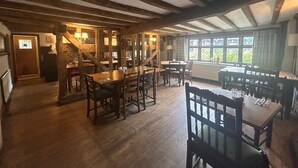 A rustic dining area with wooden tables and chairs, a staircase, and a view of the outdoors through large windows. - The Red Cow Inn (Royston)