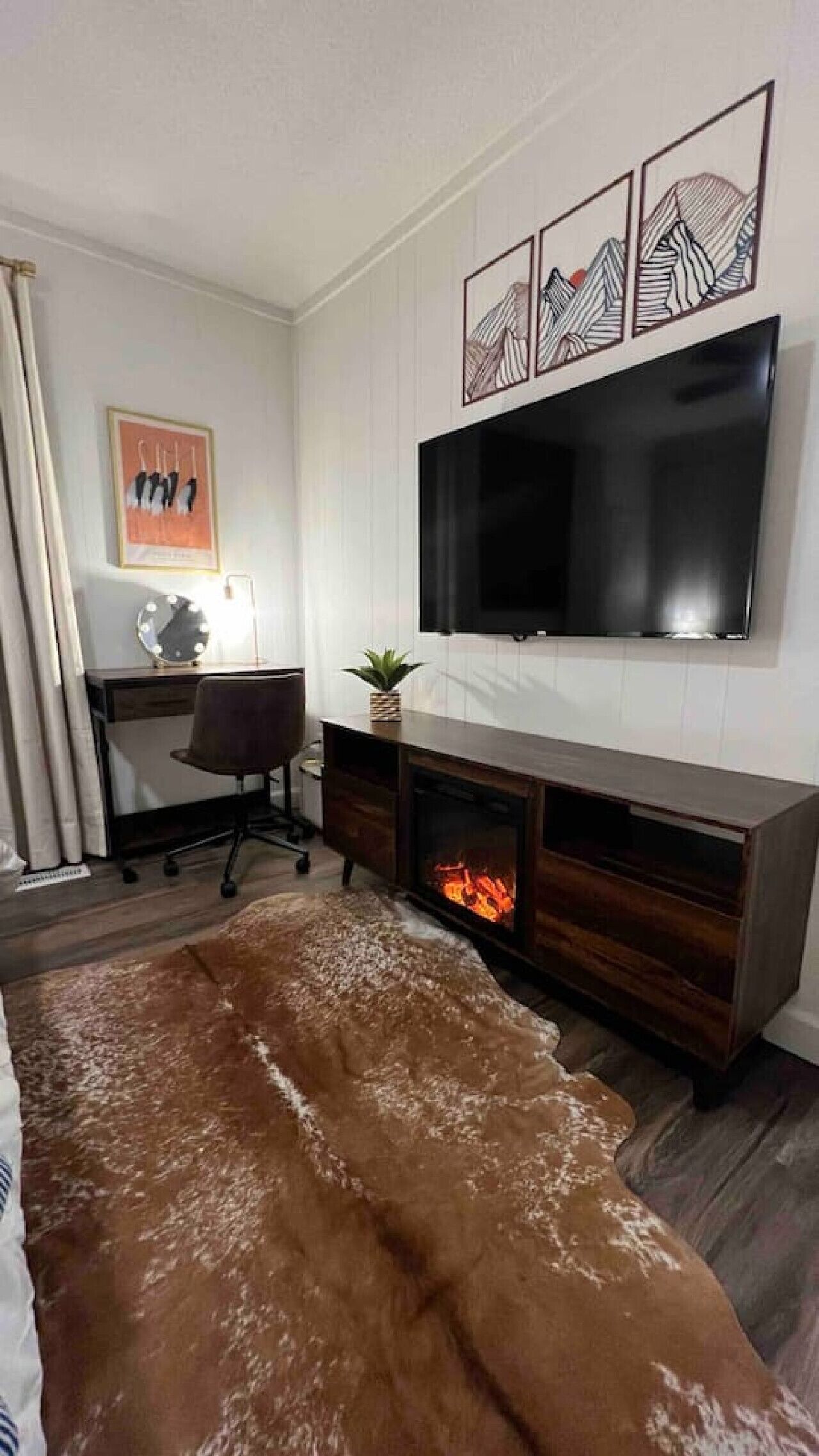 A modern living room with a flat-screen TV, a fireplace, a desk with a chair, and a cowhide rug.