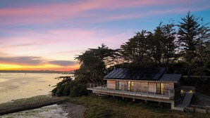 Una casa costera moderna con una terraza que da al mar, rodeada de árboles y un cielo al atardecer.