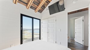 Un dormitorio con una cama grande, un televisor montado en la pared y un ventanal corredero que da a un balcón con vistas a la playa.