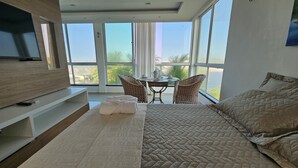 Ein modernes Schlafzimmer mit einem großen Bett, einem Fernseher auf einem Holzbodenregal, einem Esstisch mit Stühlen und einem Balkon mit Blick.