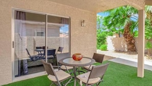 A covered patio with a round table and chairs, a decorative vase, and a view of a palm tree and greenery.