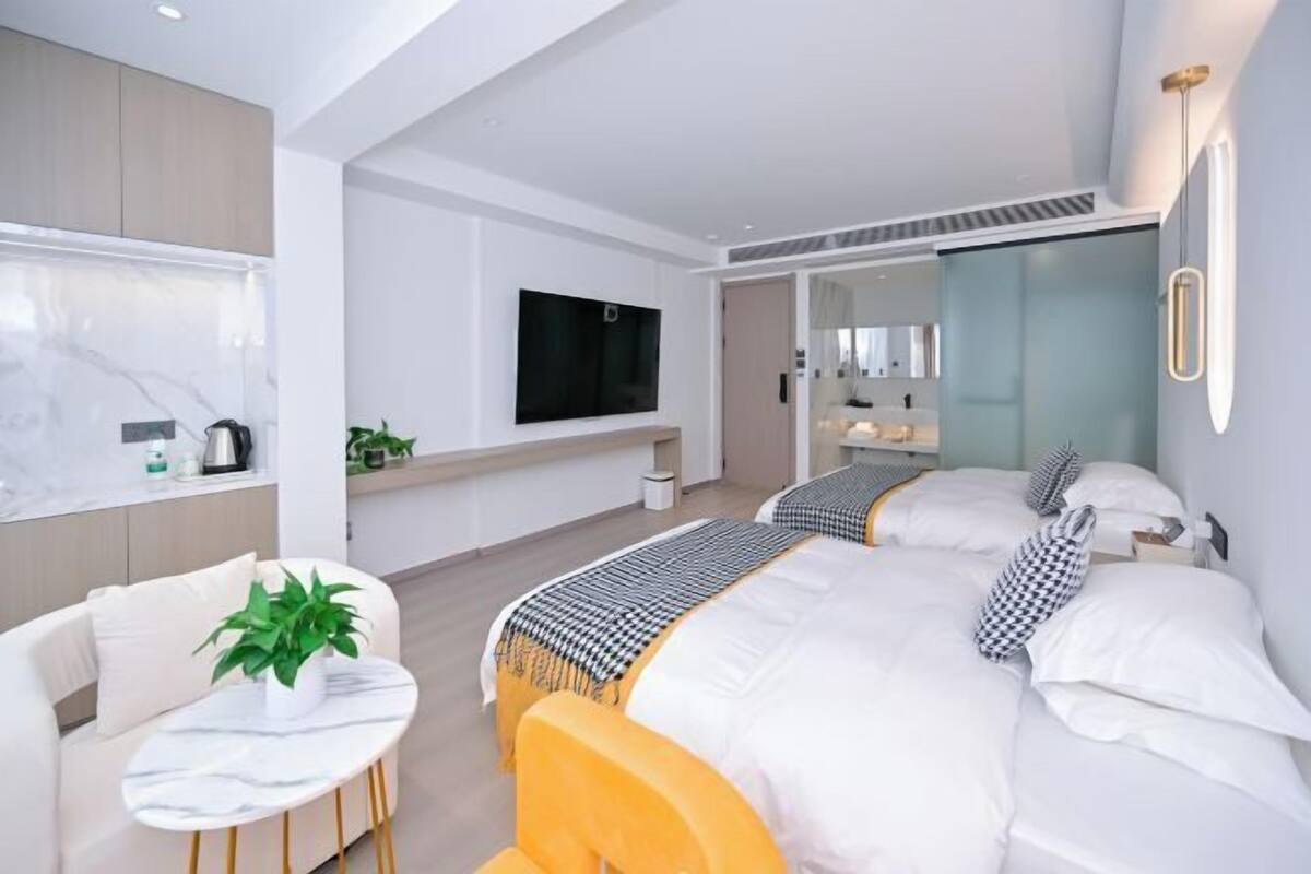 Una habitación de hotel moderna con una cama grande, un sofá, una mesita con una planta, un televisor y un baño visible a través de una puerta de cristal.