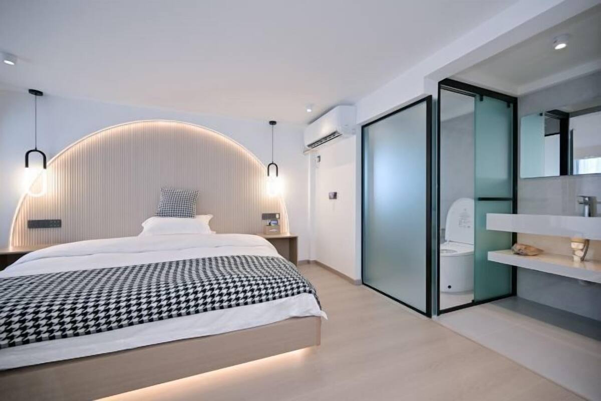 Una habitación de hotel moderna con un cabecero curvo, una cama con edredón a cuadros, una mesita de noche con lámpara, un separador de vidrio que divide el espacio y un baño con inodoro y lavamanos.