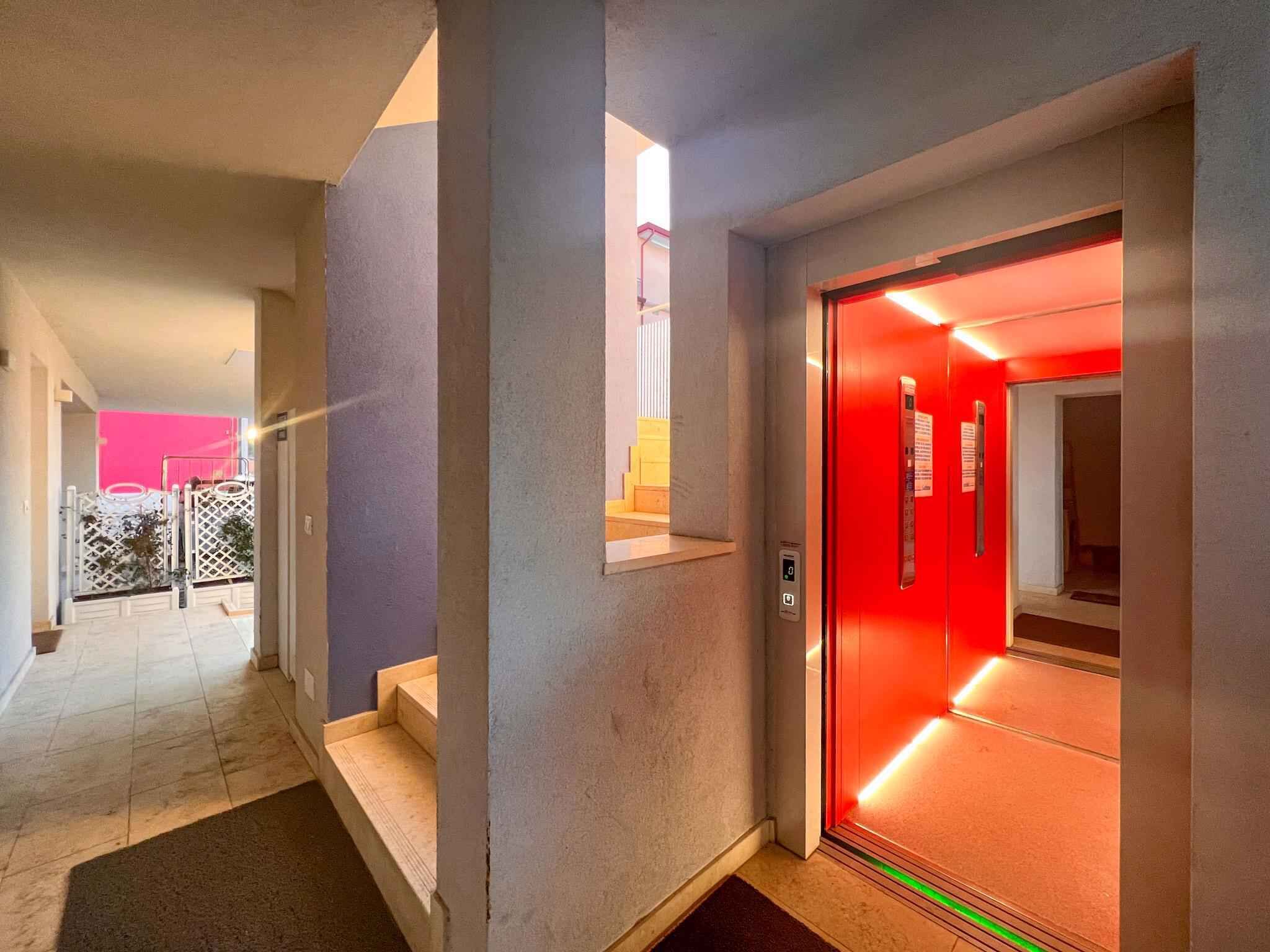 Un couloir avec un ascenseur rouge, un escalier et une porte donnant sur une pièce dont un mur est rose.