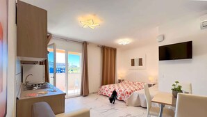 Ein Hotelzimmer mit Küchenzeile, einem Bett mit floralem Überwurf, einem an der Wand befestigten Fernseher und einem Balkon mit Blick auf Gebäude.