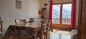 A dining area with a table covered in a patterned tablecloth, wooden chairs, a red curtain, and a view of snow-covered mountains through a window.