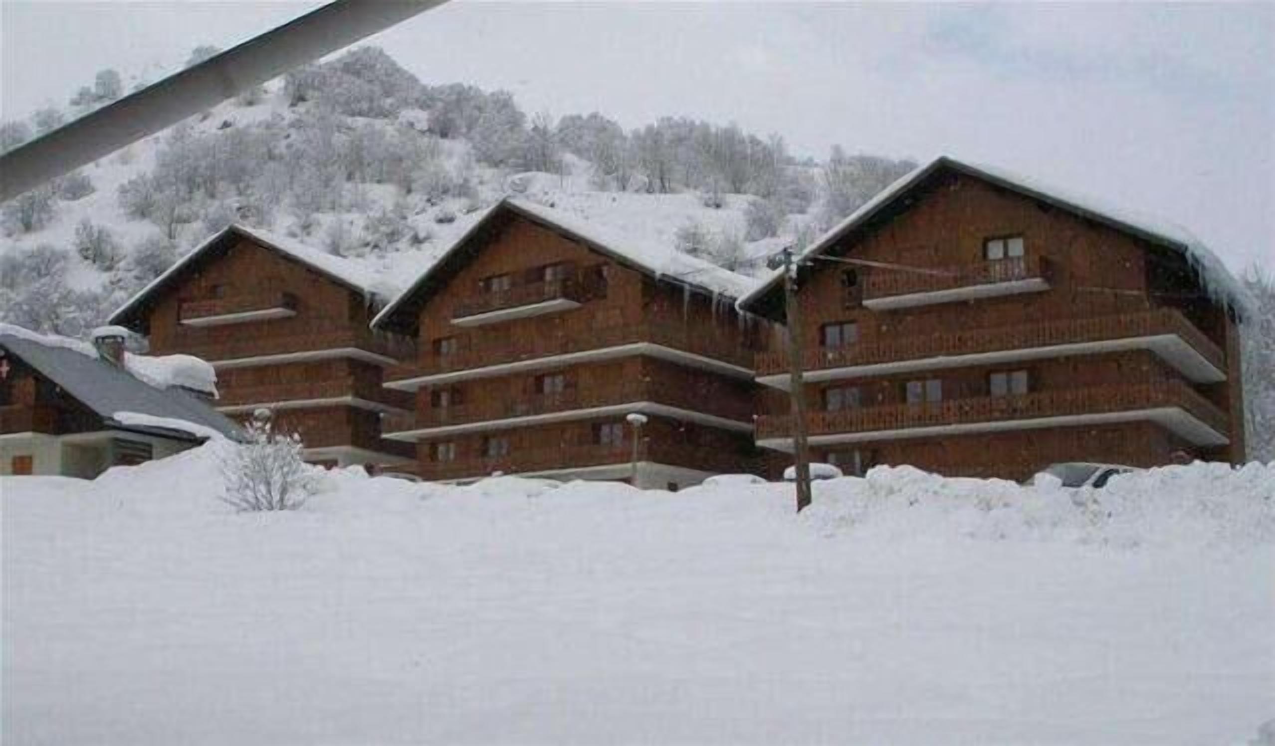 Una hilera de cabañas de madera con balcones, rodeadas por terreno nevado y una colina boscosa al fondo.