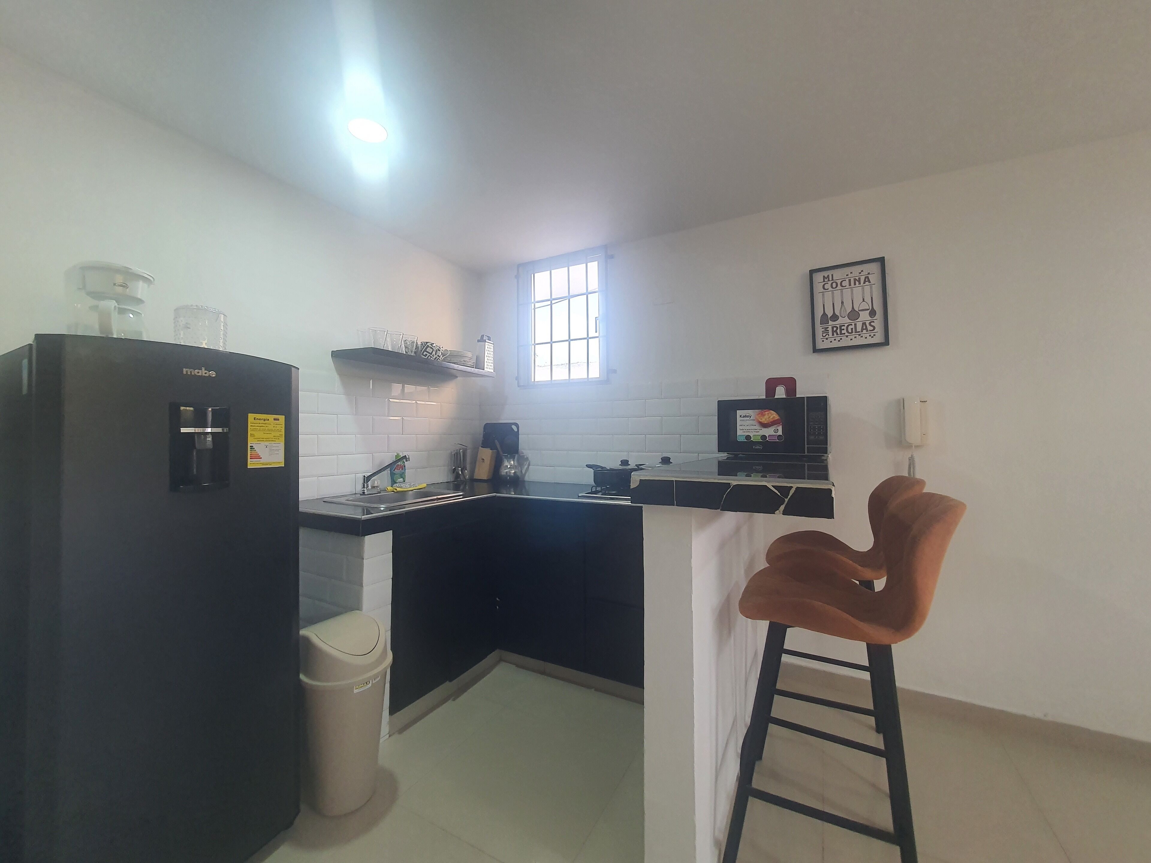 A compact kitchen with a refrigerator, sink, and a microwave. There's a window providing natural light and a small stool near the counter.