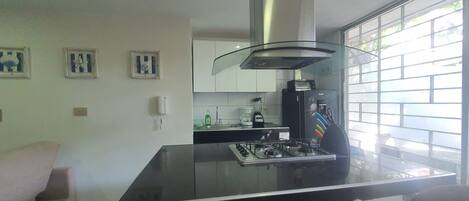 Eine moderne Küche mit einem Gasherd, einer Mikrowelle und einer Kaffeemaschine. Im Wohnbereich befindet sich ein Fenster mit Jalousien, eine weiße Wand und ein rosa Sofa.