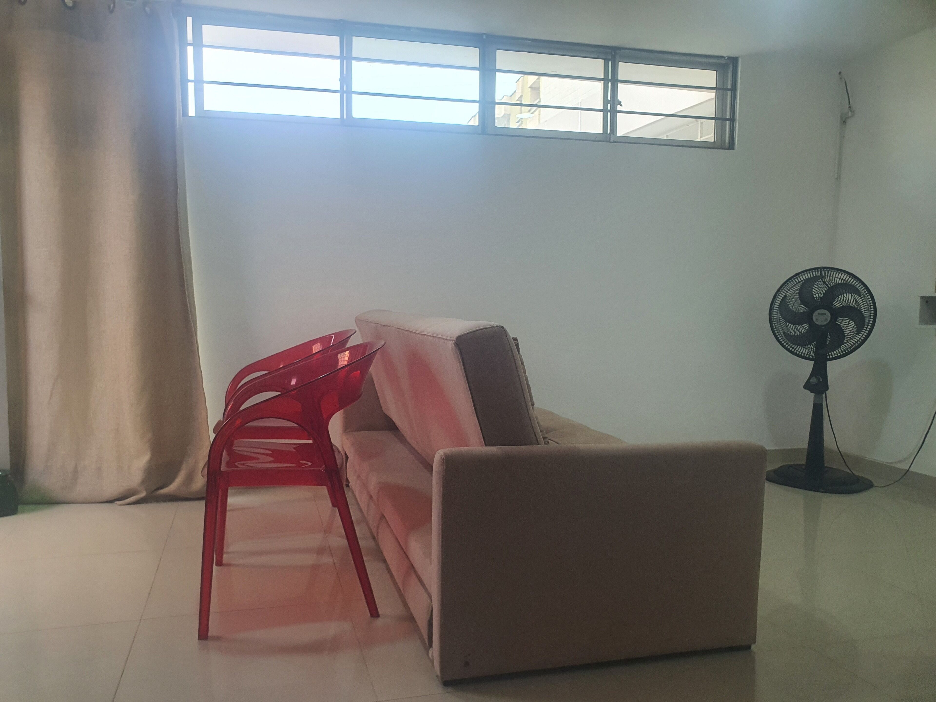 A room with a beige sofa, a red chair, a standing fan, and a window with curtains.