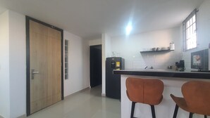 A modern kitchen with a black countertop, two orange bar stools, a coffee machine, and a refrigerator. - Casa Esther Apartaestudios (Barranquilla)