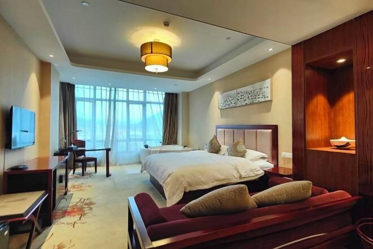 A hotel room with a bed, a desk, a TV, a large window, and a ceiling light.