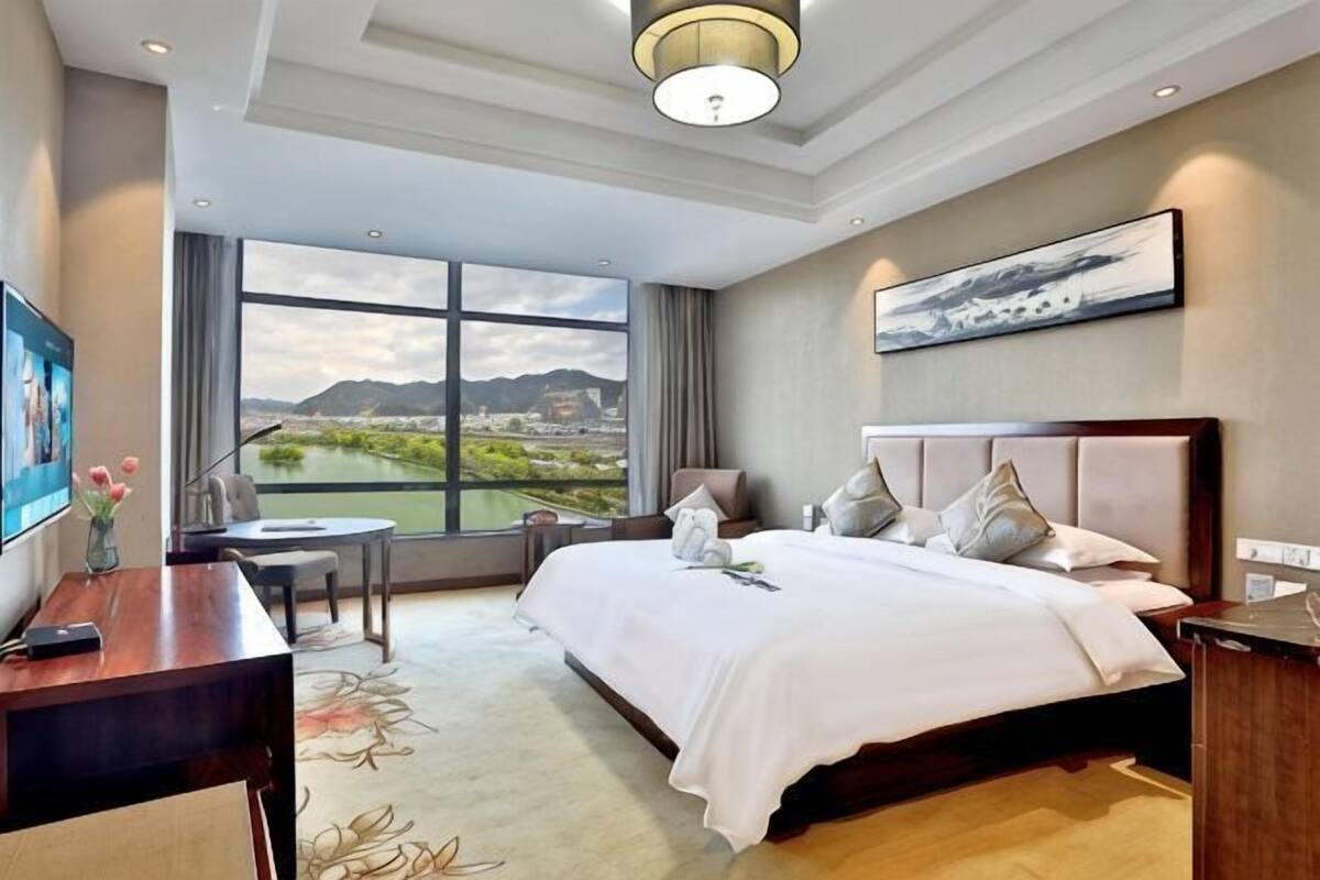 A hotel room with a large bed, a desk, a chair, a small table, and a view of a landscape through large windows.