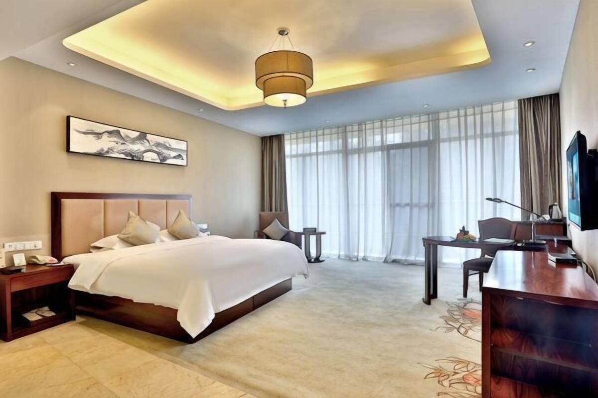 A hotel room with a large bed, a desk, a chair, a TV, and a large window with curtains.