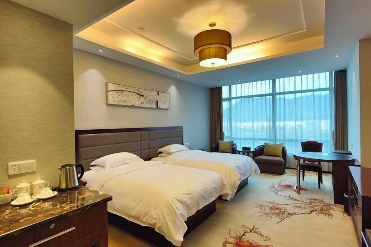 A hotel room with two beds, a desk, a chair, a coffee maker, and a large window with curtains.