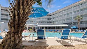 A pool area with lounge chairs and umbrellas, a palm tree, and a multi-story building with balconies in the background. - Island Sunrise HHI Condo (Hilton Head Island)