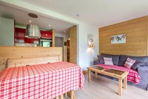 A room with a bed, a sofa, a wooden table, and a kitchenette in the background.