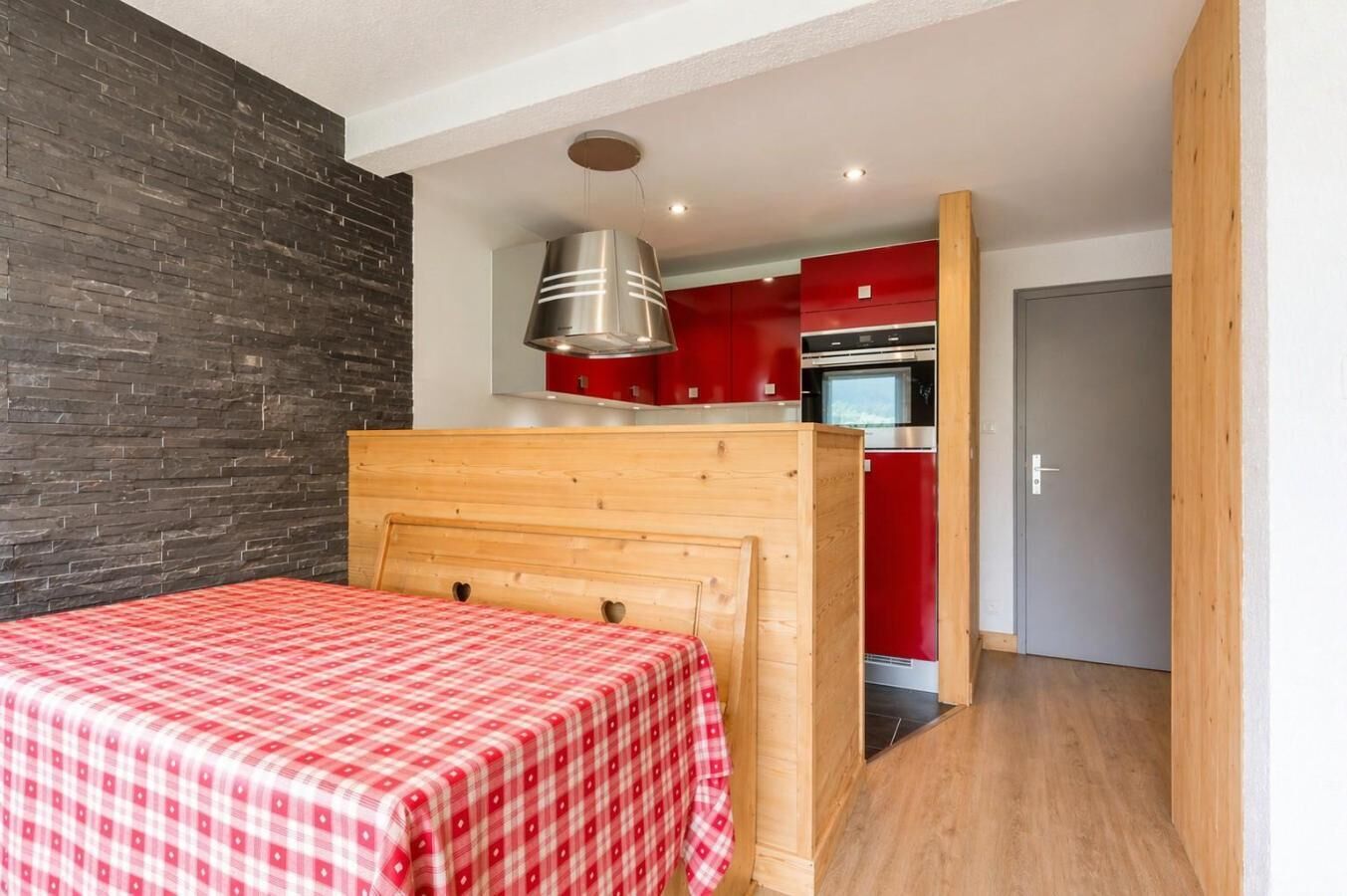 A bedroom with a wooden headboard, a red checkered bedspread, a modern kitchen with red cabinets, and a grey door.
