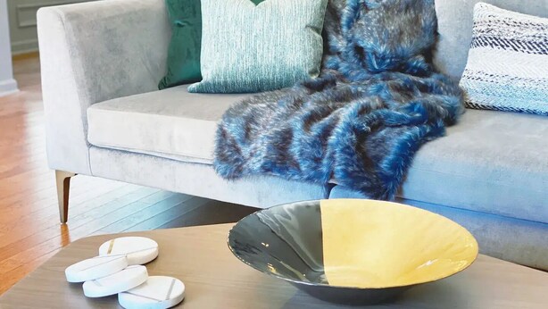 Una sala de estar moderna con un sofá gris, una manta azul y gris, y una mesa de centro de madera con un cuenco negro y amarillo y posavasos blancos.