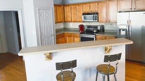 Una cocina con armarios de madera, un refrigerador de acero inoxidable, un microondas, una estufa y dos taburetes de bar.