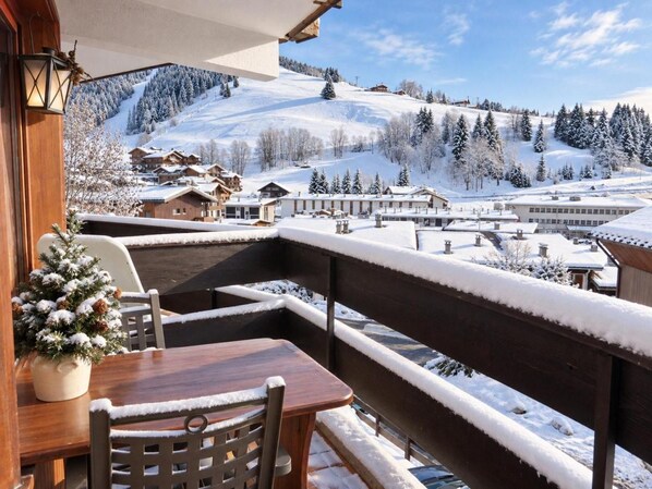 A snow-covered balcony with a wooden table and chairs, overlooking a snowy village and mountains.