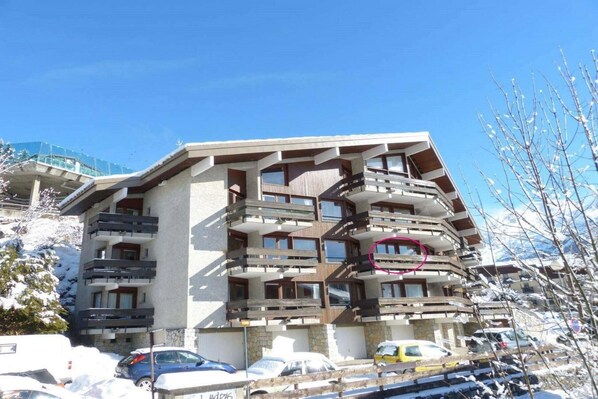 A multi-story hotel with balconies, a parking area, and a snowy mountain backdrop.