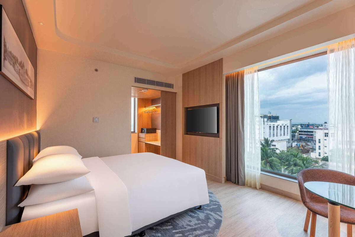 A hotel room with a bed, a TV mounted on the wall, a window with curtains, and a view of the cityscape.