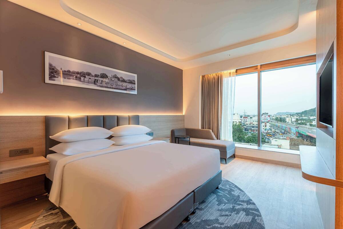 A modern hotel room with a large bed, a flat-screen TV, a sofa, and a city view.