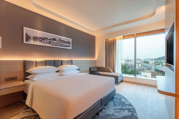 A modern hotel room with a large bed, a flat-screen TV, a sofa, and a city view.