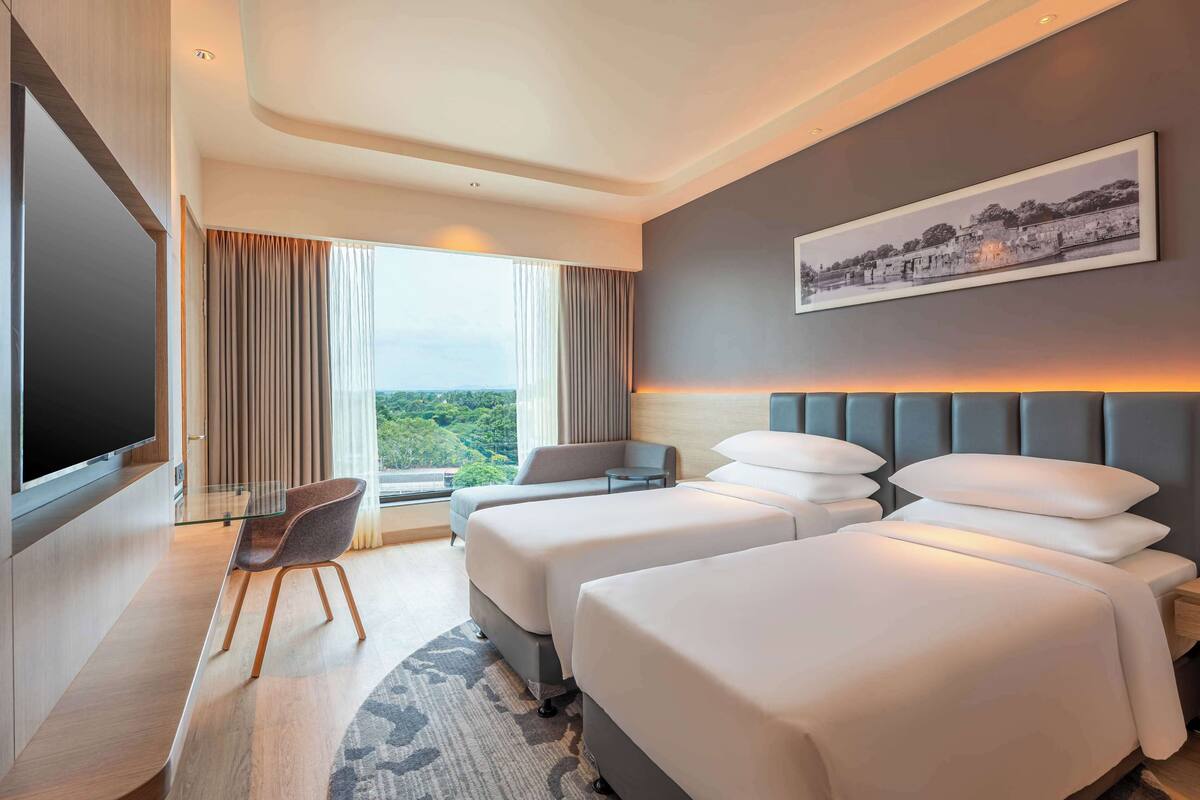 A hotel room with two beds, a TV, a desk, and a large window with curtains.
