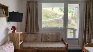Ein gemütliches Zimmer mit Schlafsofa, einem kleinen Holztisch, einem an der Wand befestigten Fernseher und einem Fenster mit Blick auf eine grüne Landschaft.