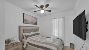 Une chambre à coucher comprenant un lit, une table de chevet, une lampe, un ventilateur de plafond, une télévision et des rideaux.