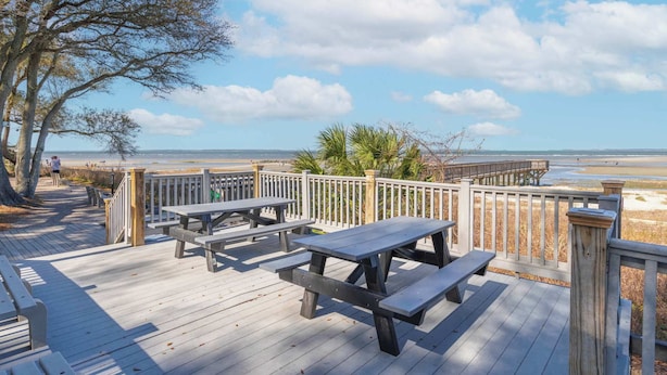 Une terrasse en bois avec des tables de pique-nique et une rambarde en bois donnant sur une plage.