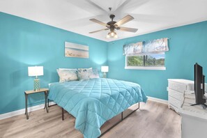 A bedroom with teal walls, a bed with a blue quilt, two bedside tables with lamps, a ceiling fan, a window with a view, and a white dresser. - Beachy Vibe – 2 BDR In Desirable HHI Resort! (Hilton Head Island)