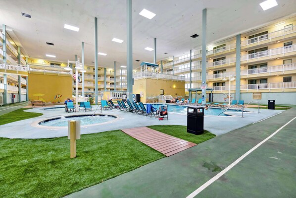 An indoor swimming pool with multiple levels of balconies, a central pool area with lounge chairs, and a grassy area. - Beachy Vibe – 2 BDR In Desirable HHI Resort! (Hilton Head Island)