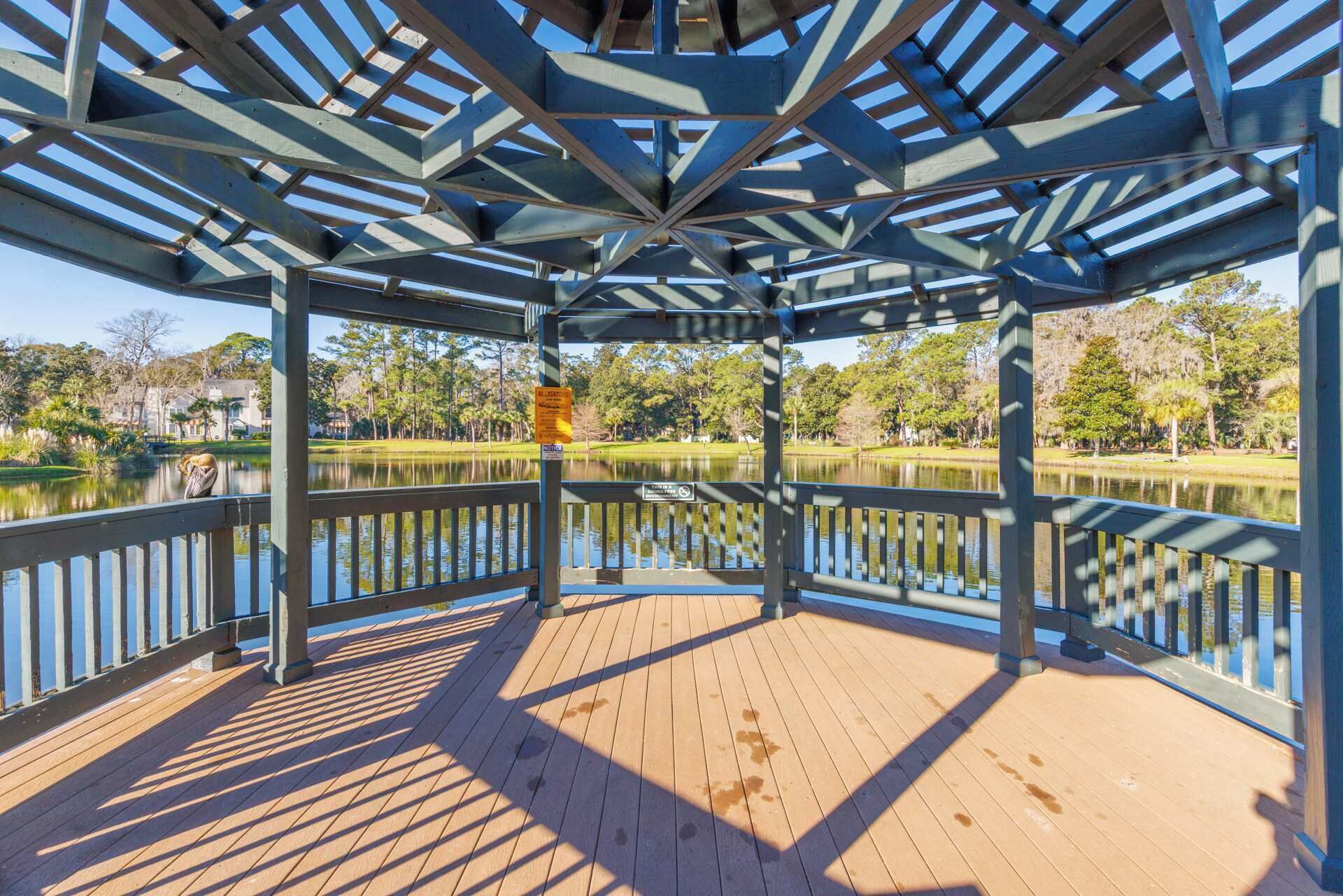 Una terraza cubierta de madera con estructura metálica, con vista a un lago tranquilo y a un parque con árboles y un edificio al fondo.