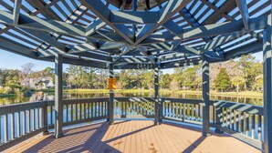 Una terraza cubierta de madera con estructura metálica, con vista a un lago tranquilo y a un parque con árboles y un edificio al fondo.