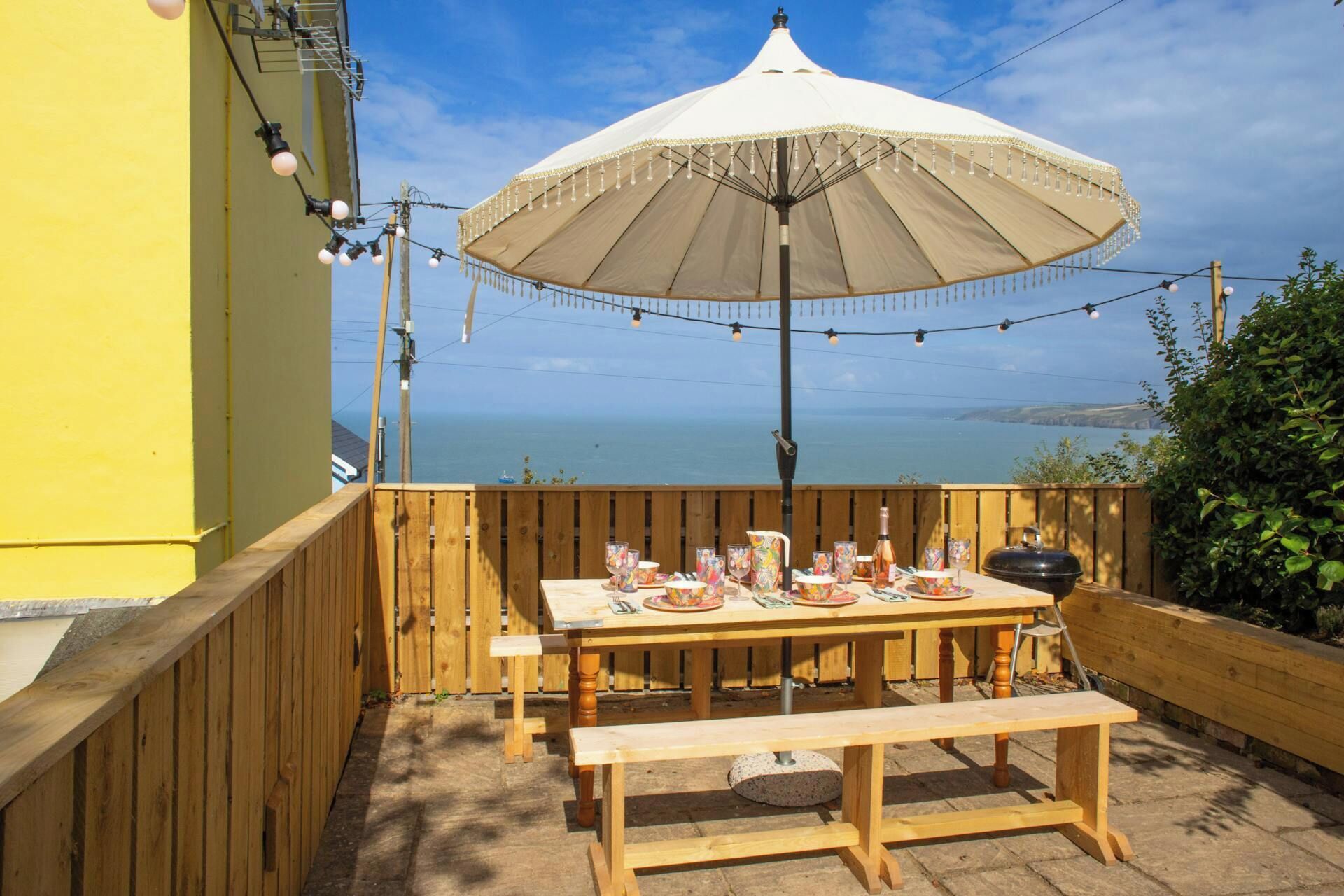 An outdoor dining area with a table set for four, a large umbrella, and a view of the sea.