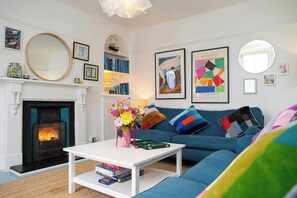 A living room with a fireplace, a sofa, a coffee table with books and a vase of flowers, and artwork on the walls.