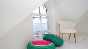 A room with a white chair, a window with a view, and two green and red cushions.