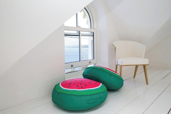 A room with a white chair, a window with a view, and two green and red cushions.