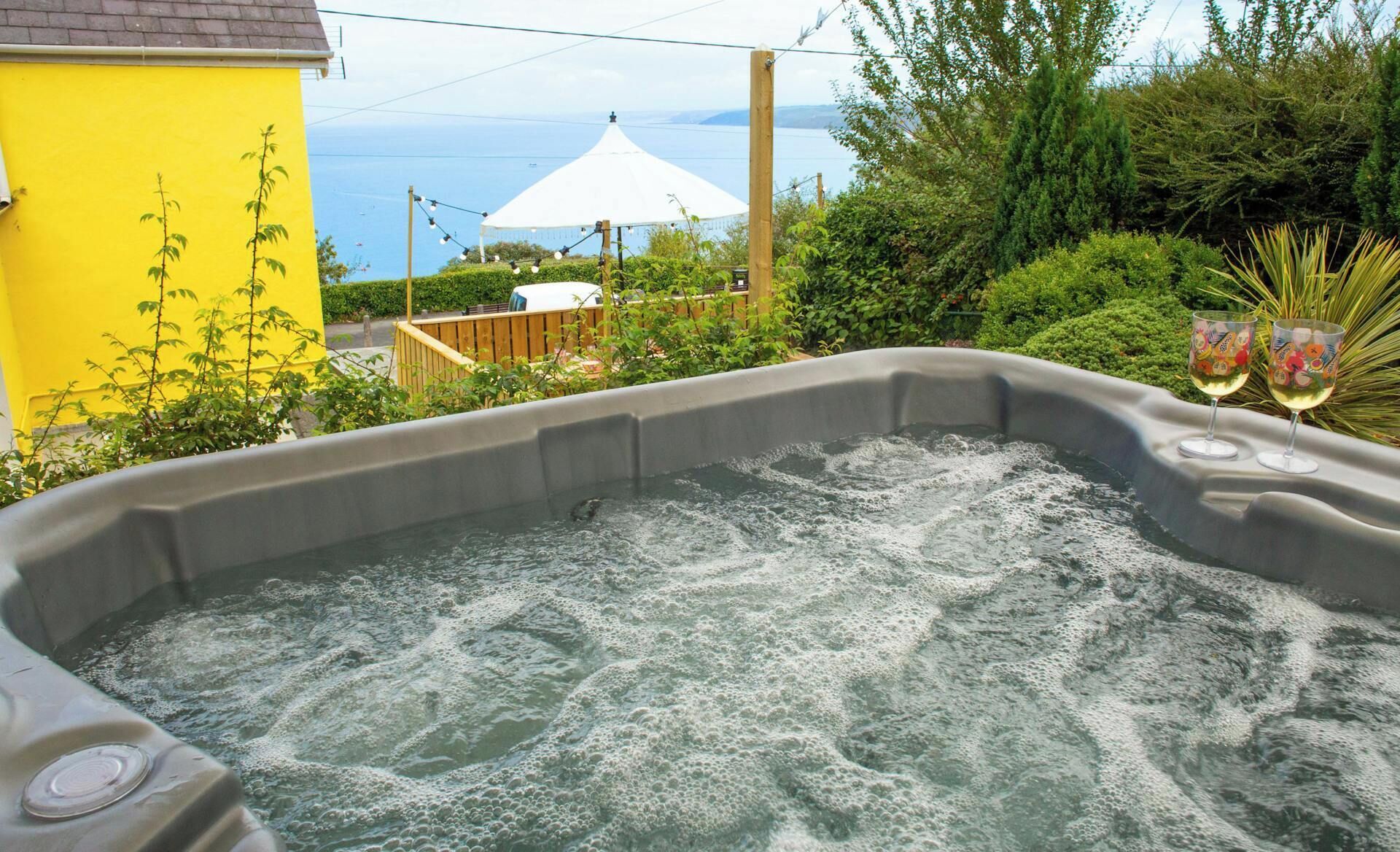 A hot tub with a view of the sea, surrounded by greenery and a yellow building.