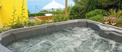 A hot tub with a view of the sea, surrounded by greenery and a yellow building.