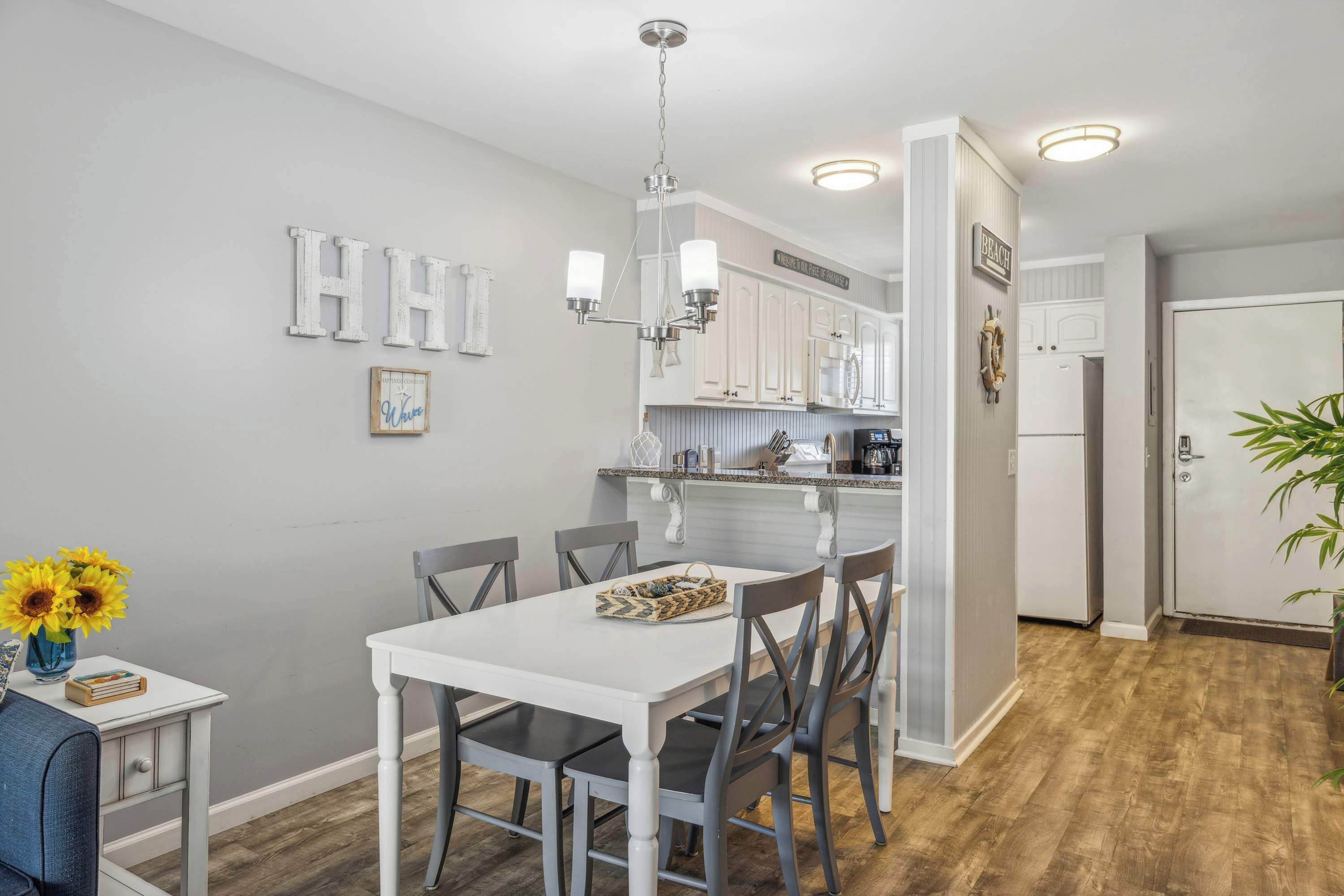 A dining area with a white table and chairs, a chandelier, and a kitchen in the background.