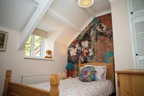 Ein Schlafzimmer mit schrägen Wänden, einem Bett mit floraler Bettwäsche, einem Teddybären auf einer Fensterbank und einem farbenfrohen abstrakten Gemälde an der Wand.