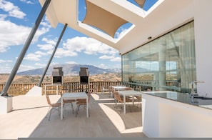 A modern outdoor dining area with a view of mountains, a glass wall, and a barbecue grill. - Overlook Apartment in Mijas - Ref 254 (Mijas)