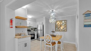 A kitchen with white cabinets, a stainless steel refrigerator, a wooden dining table with white chairs, a chandelier, and a wall-mounted artwork. - Aqua Vista Retreat - 2 BDR in Spectacular Resort! (Hilton Head Island)