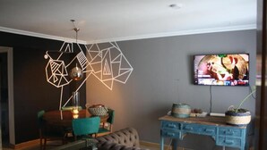 A living room with a modern geometric light fixture, a television on the wall, a blue desk, and a tufted sofa. - Charming 1-bedroom apartment with WiFi, AC in fabulous GFJ (GFJ)