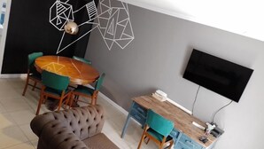 A modern living room with a round wooden dining table, green upholstered chairs, a tufted brown sofa, a flat-screen TV mounted on the wall, and a small wooden desk with a chair. - Charming 1-bedroom apartment with WiFi, AC in fabulous GFJ (GFJ)